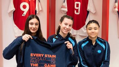 Drei Mädchen in der Umkleidekabine des Arsenal FC halten ein T-Shirt mit dem Schriftzug „Every stain should be part of the game“.